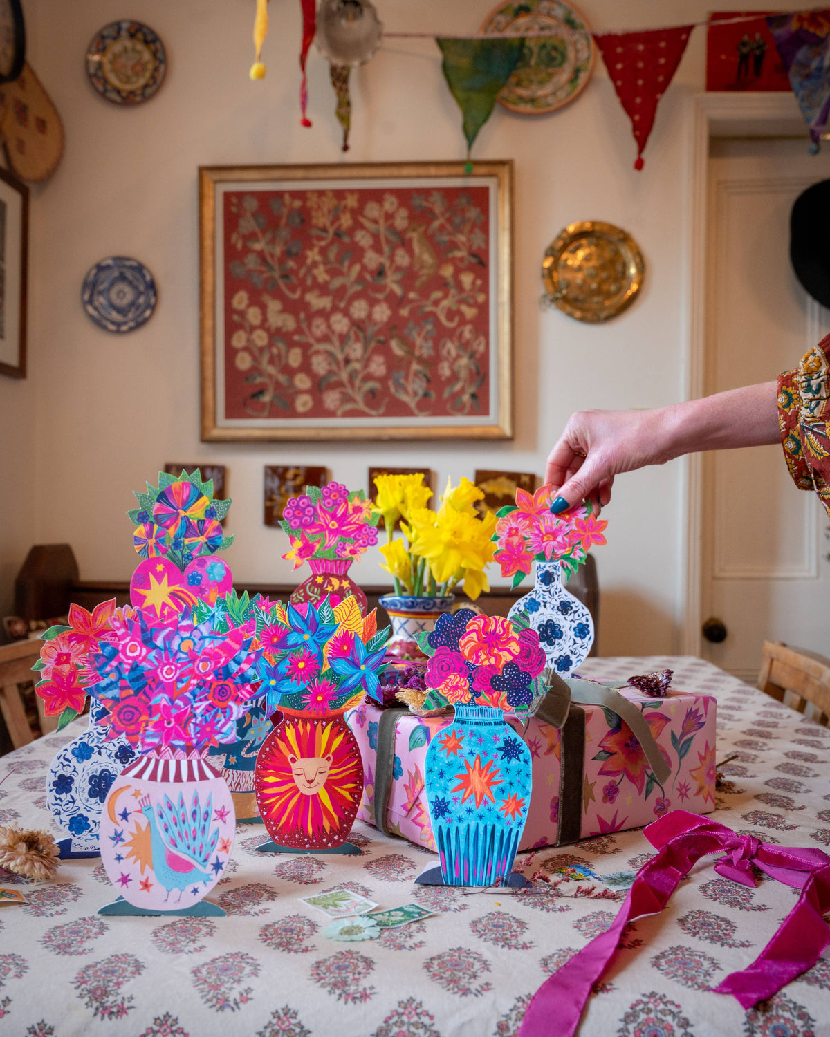 Colorful paper flower arrangements on a table with a person interacting with them, in a room with decorative plates and artwork on the wall.