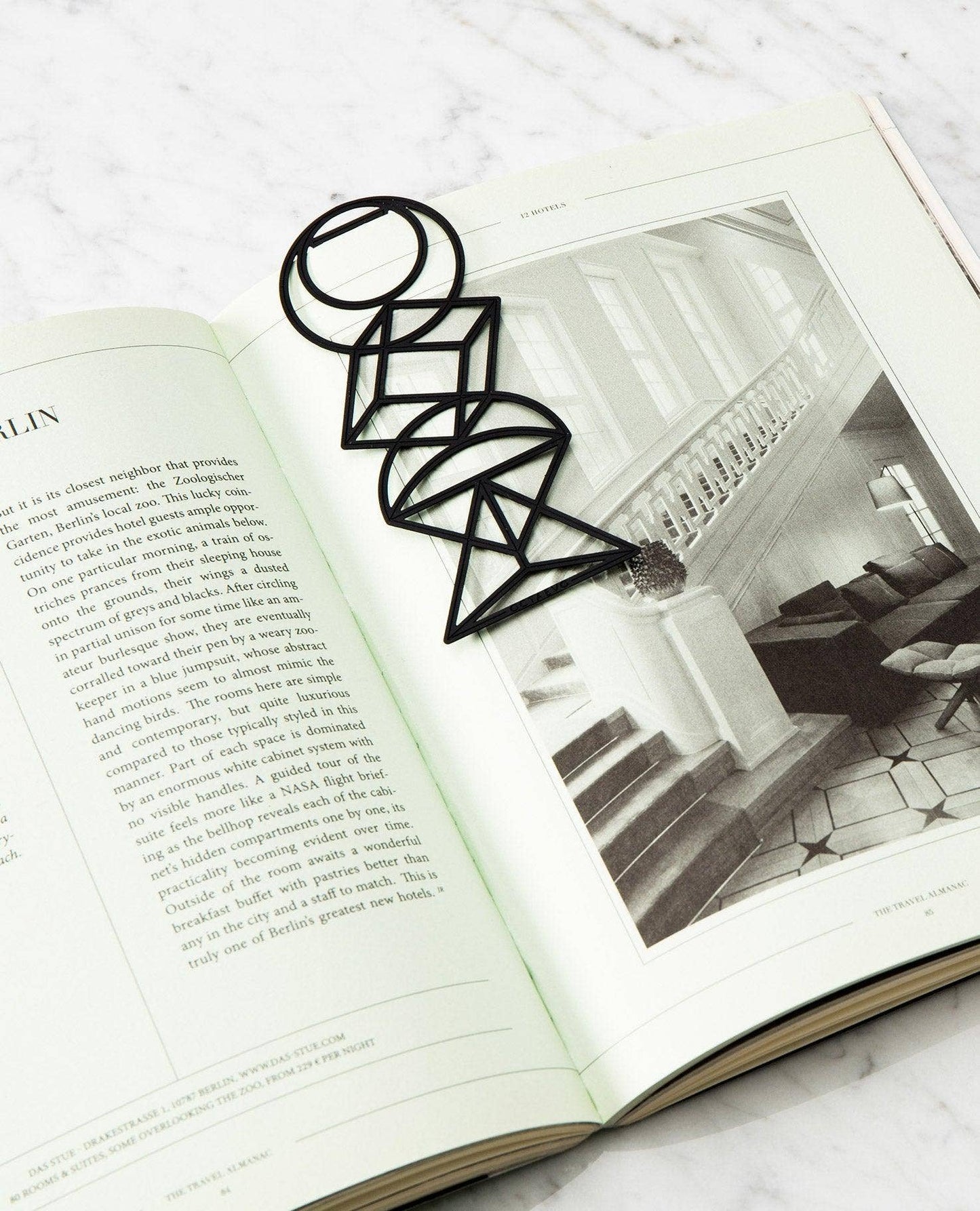 A black geometric wire bookmark shaped like a stylized letter 'O' or abstract floral design, resting on an open book with a black-and-white interior photo of a staircase.