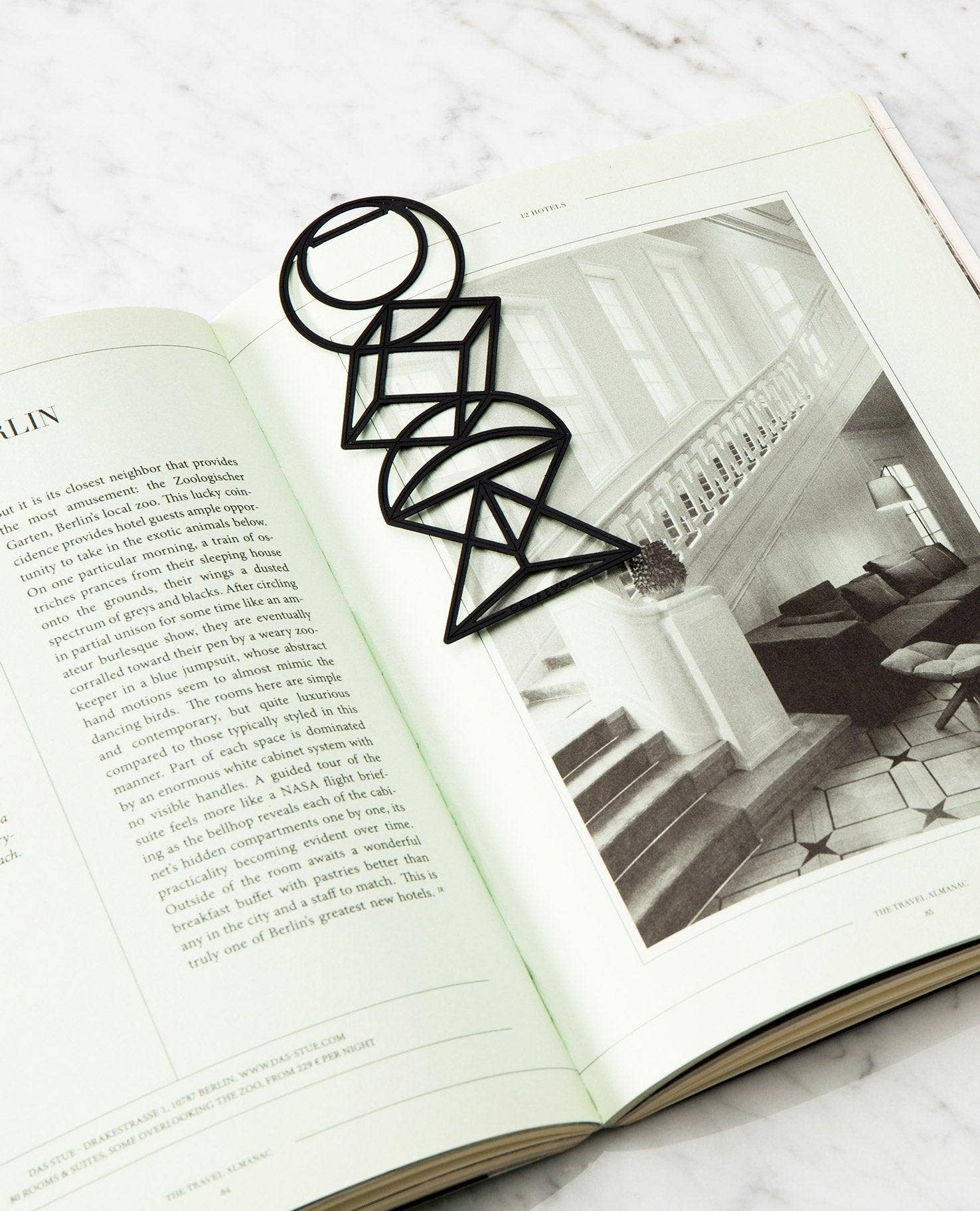 A black geometric wire bookmark shaped like a stylized letter 'O' or abstract floral design, resting on an open book with a black-and-white interior photo of a staircase.