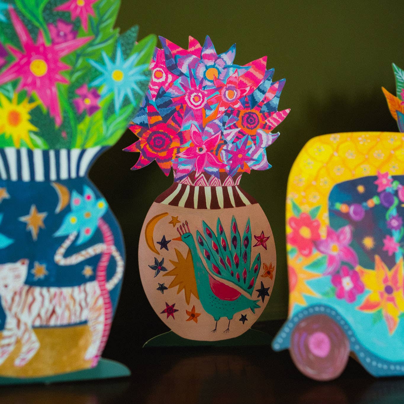 Colorful decorative items including a vase with flowers, a peacock design, and a floral-patterned box on a dark background.