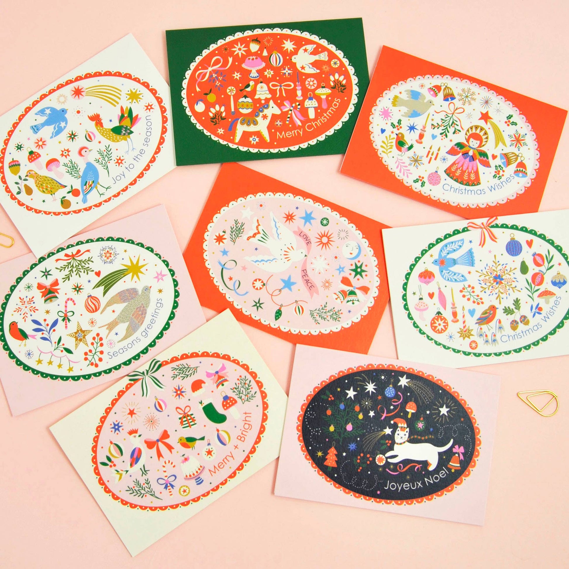 A set of colorful oval Christmas greeting cards arranged on a light pink background. The cards feature whimsical holiday illustrations, including a dog, birds, stars, ornaments, and festive scenes, with different vibrant card backings in green, orange, pink, and cream.