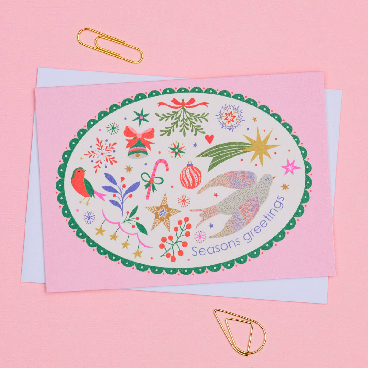 A pink Christmas greeting card with an illustrated oval design featuring birds, stars, ornaments, candy canes, and the text 'Seasons greetings,' shown with a white envelope on a pink background with gold paperclips.