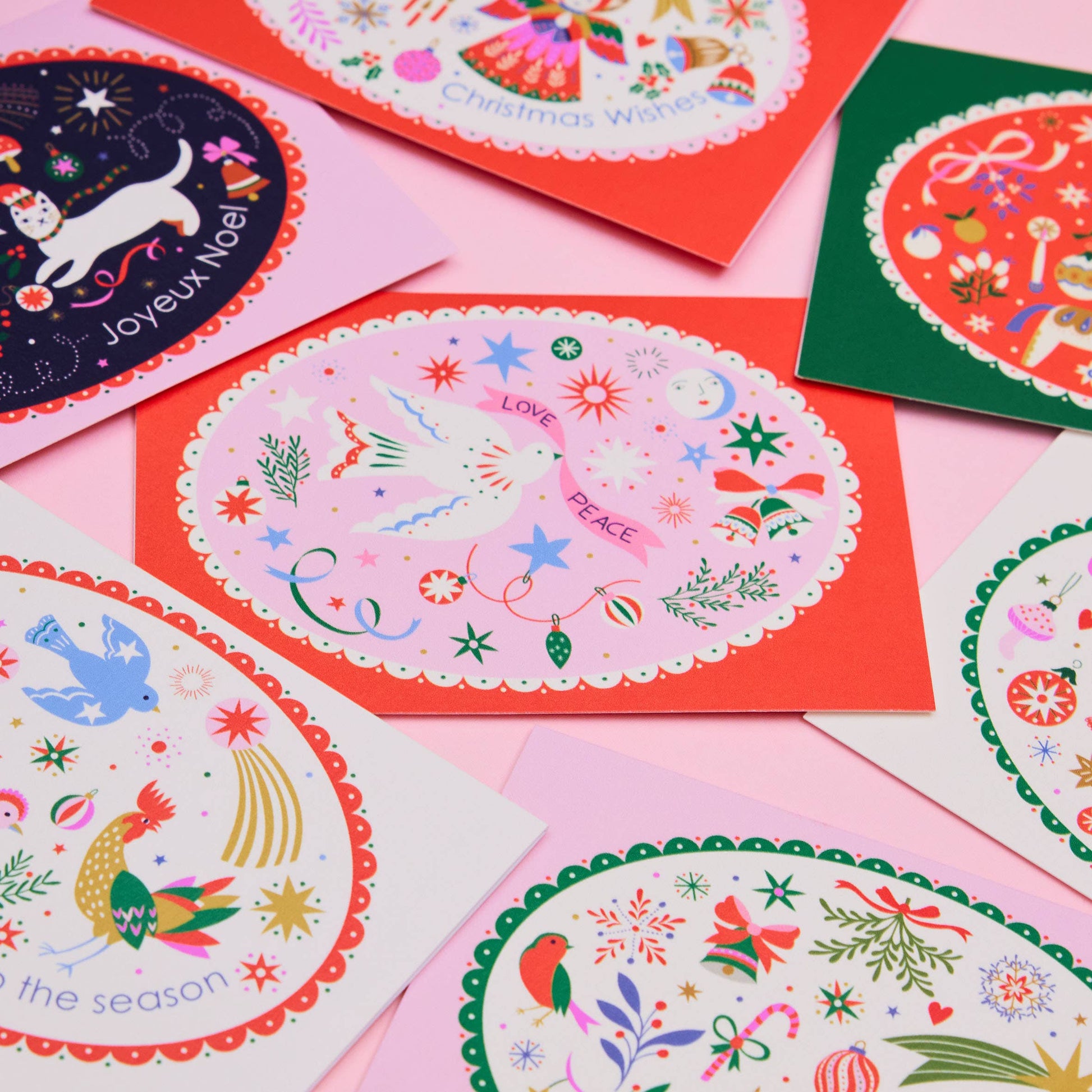 A flat-lay arrangement of multiple Christmas greeting cards with colorful holiday illustrations, including birds, snowflakes, ornaments, and festive text, set against a light pink background.