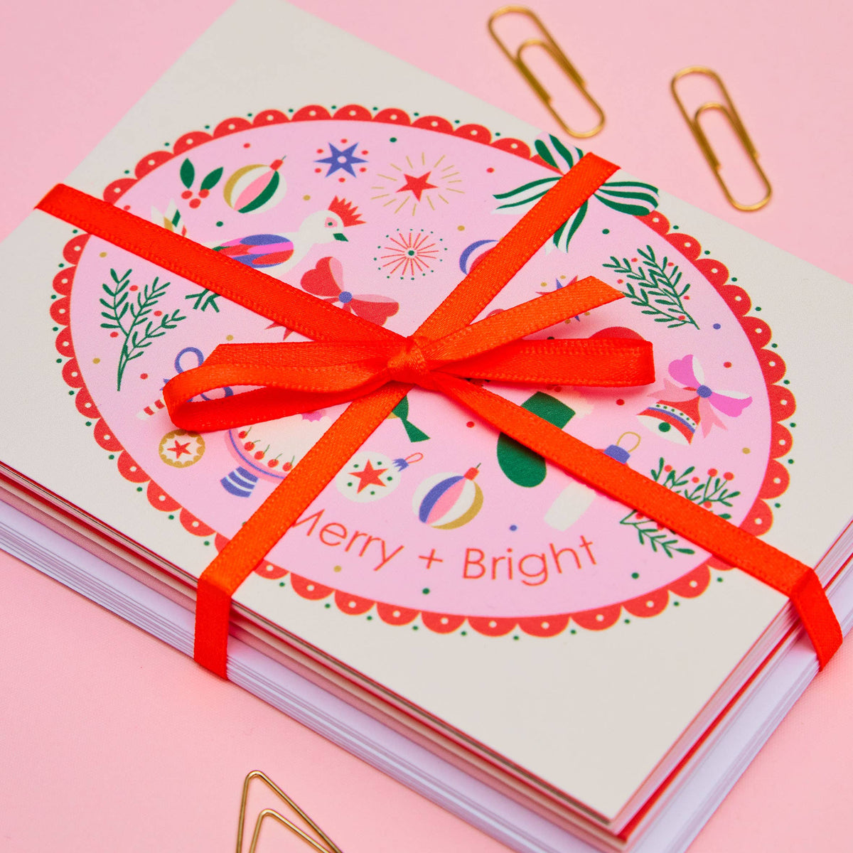 A stack of greeting cards tied with a bright orange-red ribbon, featuring a festive oval illustration with the words 'Merry + Bright' on a pink background, styled with gold paperclips on a pink surface.