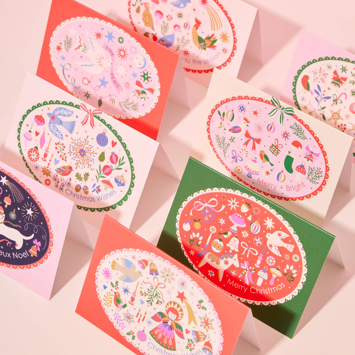 A set of Christmas greeting cards arranged on a pale pink background. The cards feature colorful oval illustrations with festive motifs such as snow globes, Christmas trees, ornaments, stars, and holiday illustrations, with phrases like 'Merry Christmas' and 'Merry Christmas Wishes'.