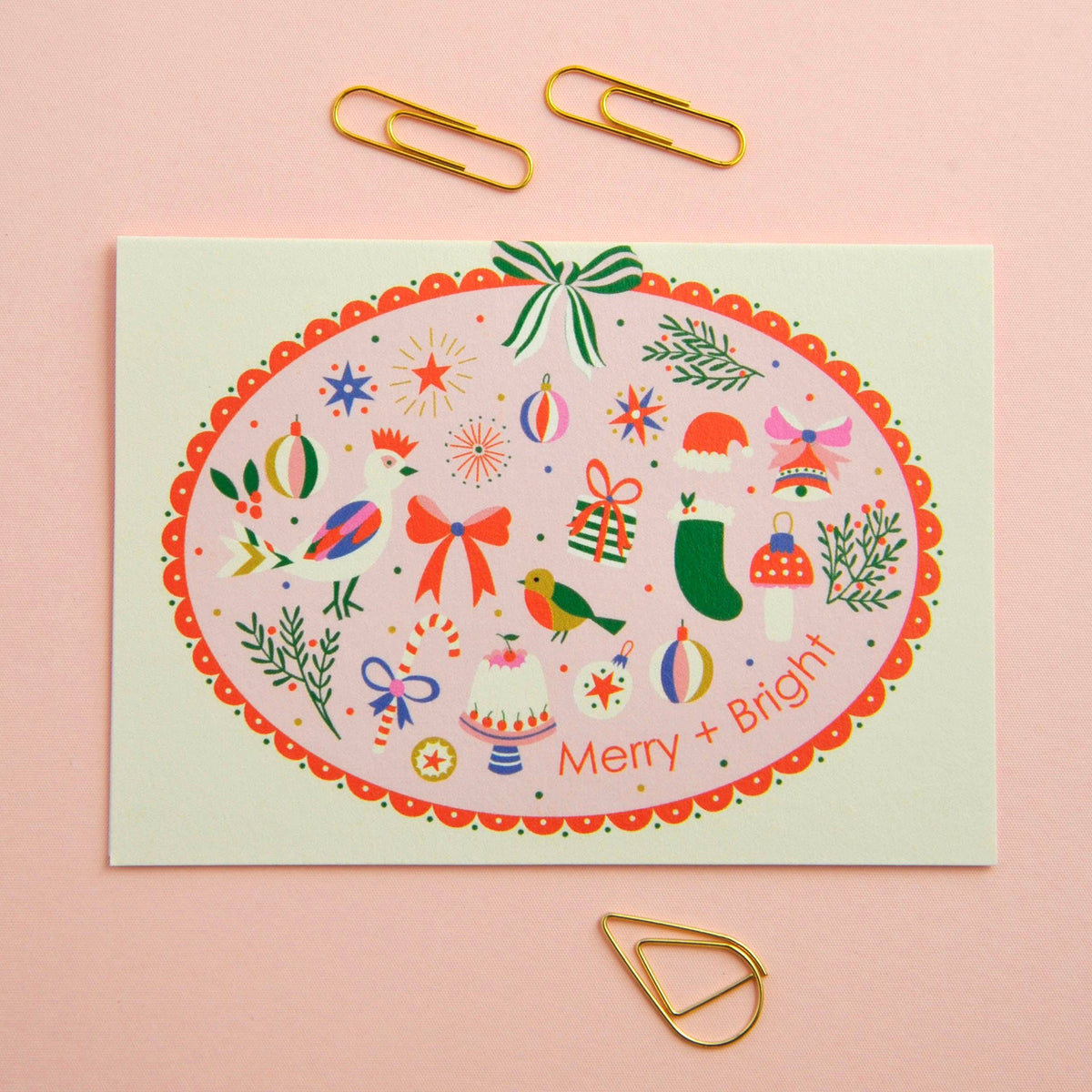 A square Christmas greeting card with a colorful oval holiday illustration and the words 'Merry + Bright' at the bottom, styled on a pink background with gold paperclips.