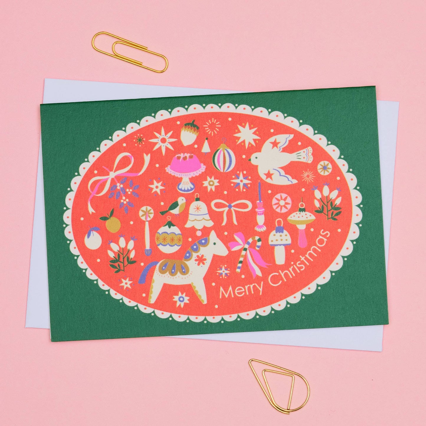 A Christmas greeting card with a dark green border and a central coral-red oval illustration featuring festive icons, a dala horse, a dove, and the words 'Merry Christmas.' The card is photographed on a pink background with gold paperclips and a white envelope visible behind it.