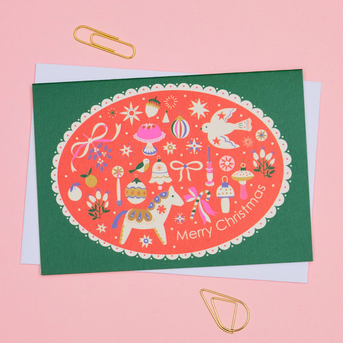 A Christmas greeting card with a dark green border and a central coral-red oval illustration featuring festive icons, a dala horse, a dove, and the words 'Merry Christmas.' The card is photographed on a pink background with gold paperclips and a white envelope visible behind it.