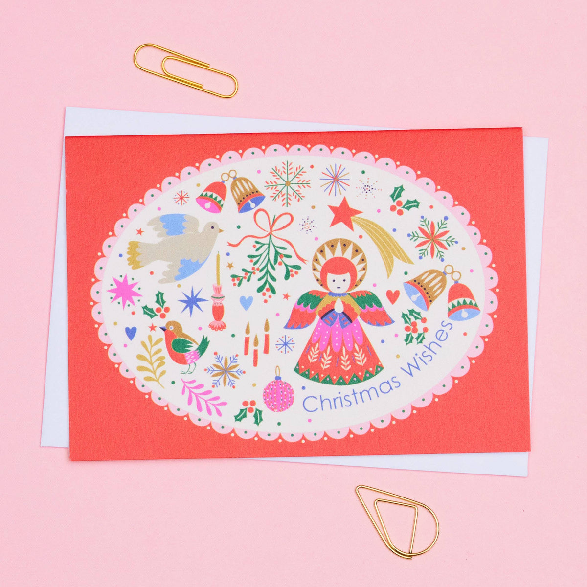 A Christmas greeting card with a coral red border and a white scalloped oval center featuring a colorful illustration of a angel, ornaments, birds, and the text 'Christmas Wishes'. The card is layered over a white envelope on a light pink background with gold paperclips.