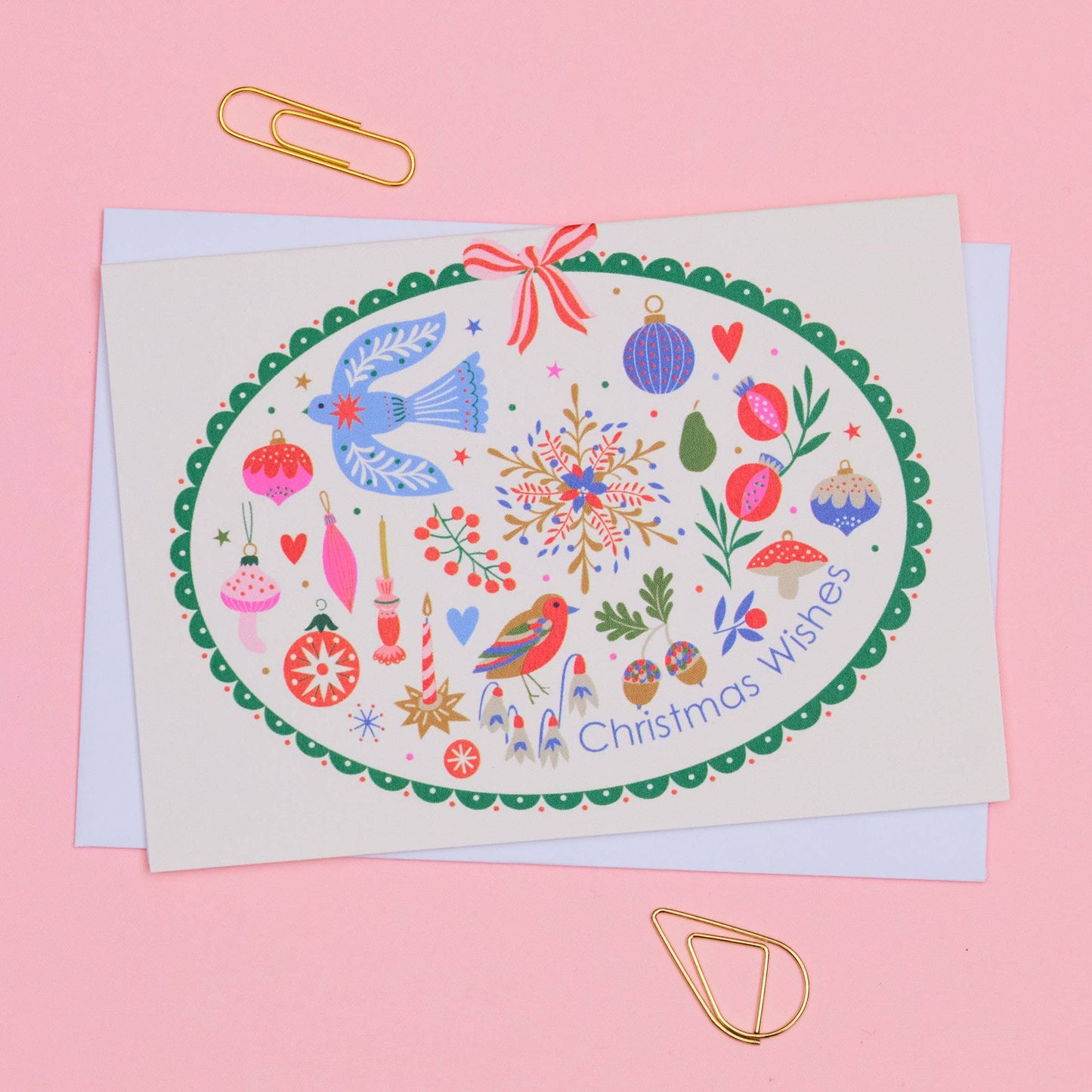 A Christmas greeting card with an oval illustration featuring birds, ornaments, snowflakes, and the text 'Christmas wishes,' photographed on a pink background with gold paperclips.