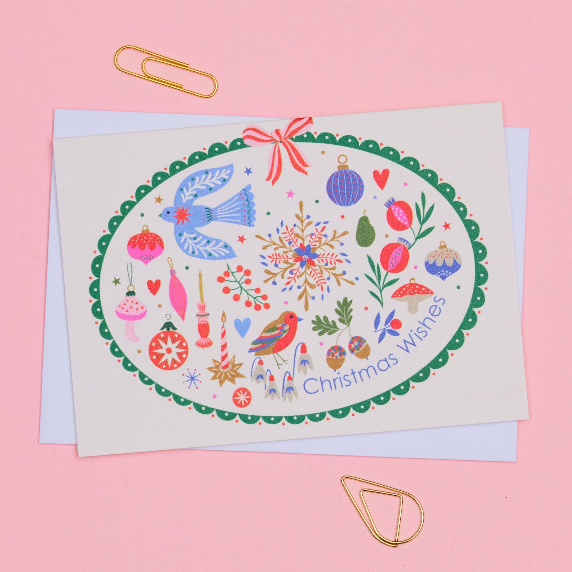 A Christmas greeting card with an oval illustration featuring birds, ornaments, snowflakes, and the text 'Christmas wishes,' photographed on a pink background with gold paperclips.