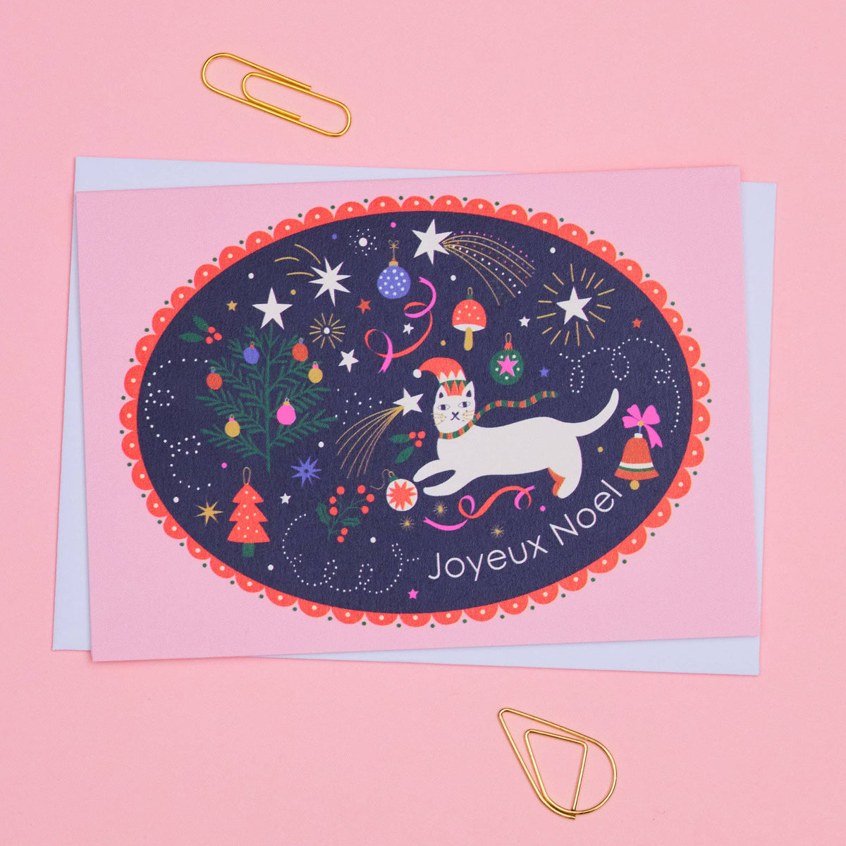 A pink greeting card with a dark navy oval illustration featuring a white cat, fireworks, stars, a Christmas tree, mushrooms, and the text 'Joyeux Noel', photographed on a pink background with gold paperclips.