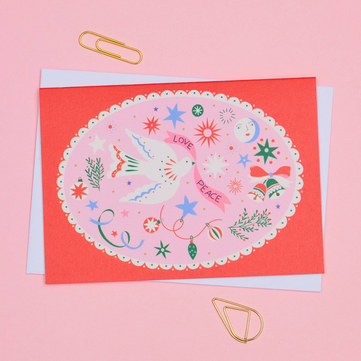 A Christmas greeting card with a coral red border and a pink center illustration featuring a white dove, stars, ornaments, and the words 'LOVE PEACE', shown with a white envelope on a pink background with gold paperclips.