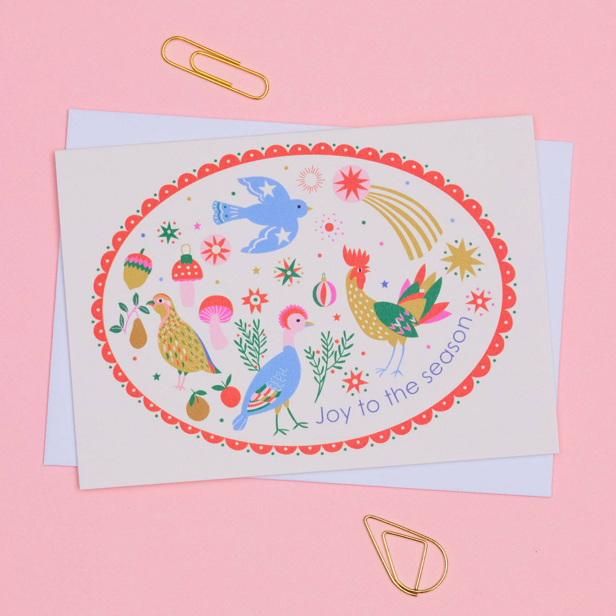A square greeting card with a colorful oval illustration of birds, flowers, and the text 'Joy to the season,' photographed on a pink background with gold paperclips and a light blue backing card visible behind it.