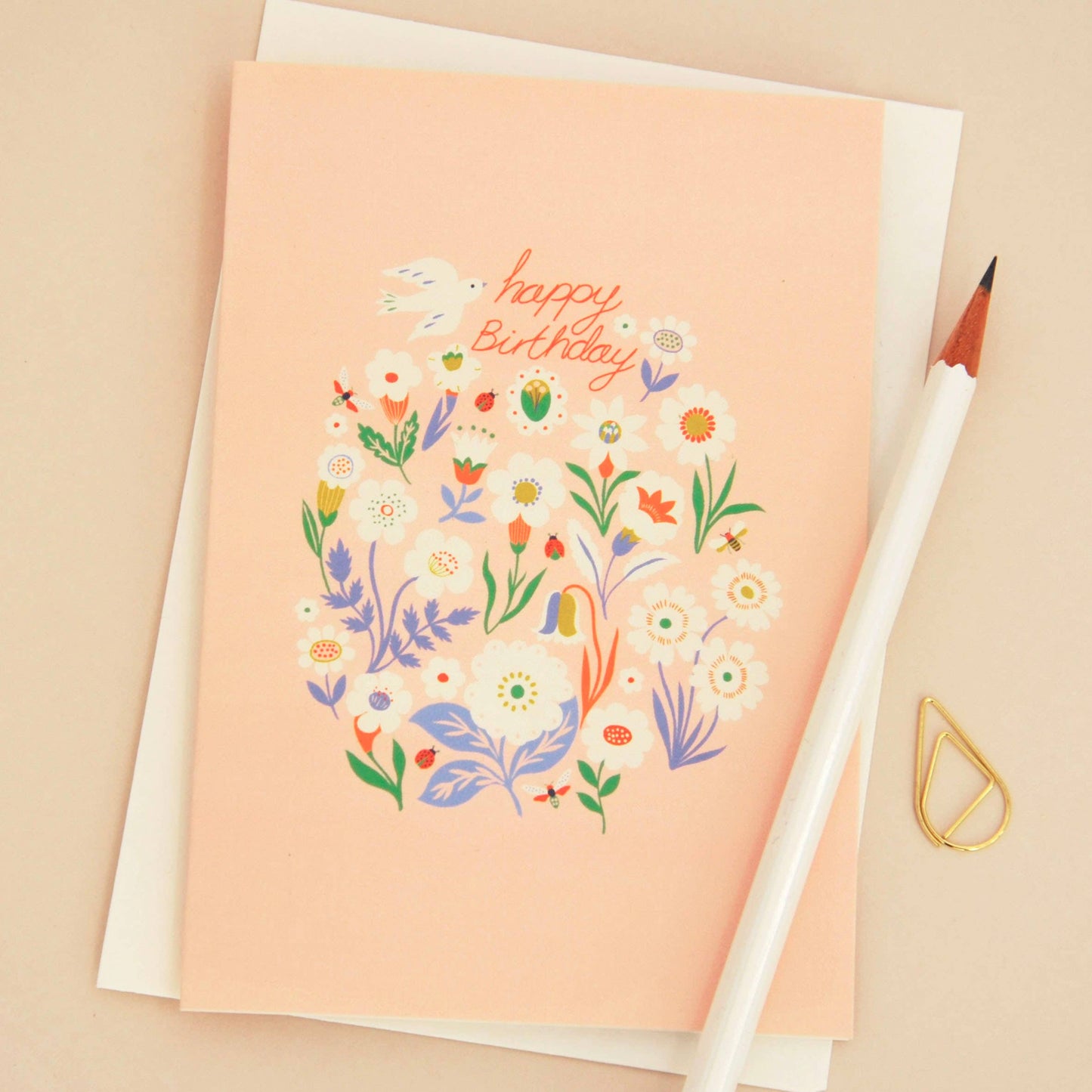 A peach-colored birthday card with a colorful floral illustration and the words 'Happy Birthday' in orange script, shown with a white envelope, a white pencil, and a small gold paperclip on a beige background.