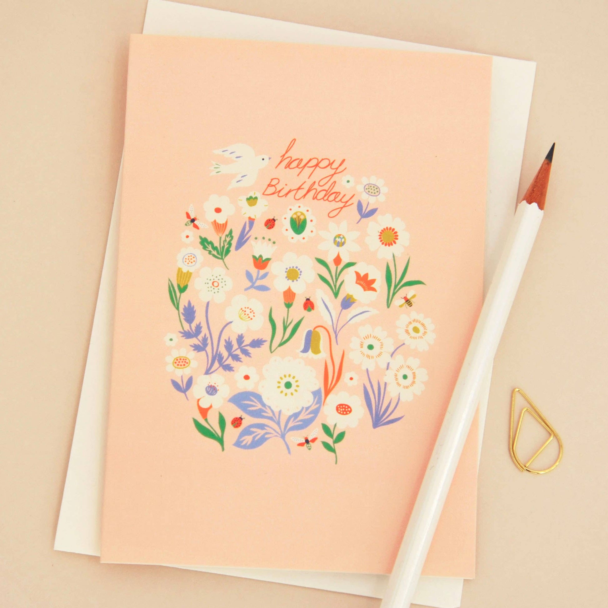A peach-colored birthday card with a colorful floral illustration and the words 'Happy Birthday' in orange script, shown with a white envelope, a white pencil, and a small gold paperclip on a beige background.