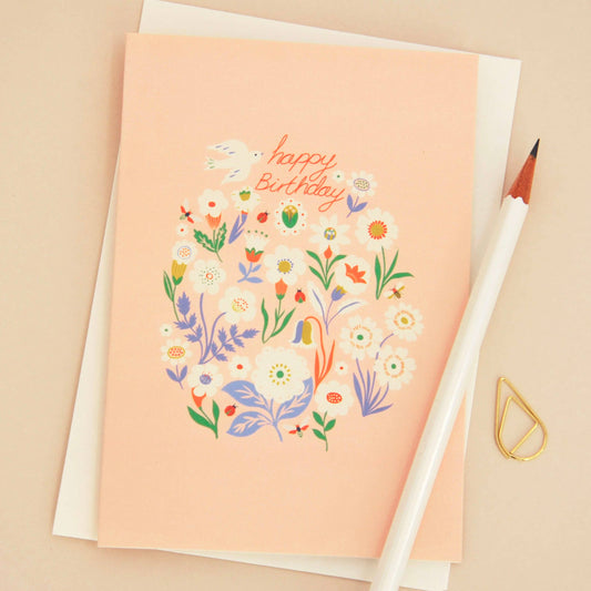 A peach-colored birthday card with a colorful floral illustration and the words 'Happy Birthday' in orange script, shown with a white envelope, a white pencil, and a small gold paperclip on a beige background.
