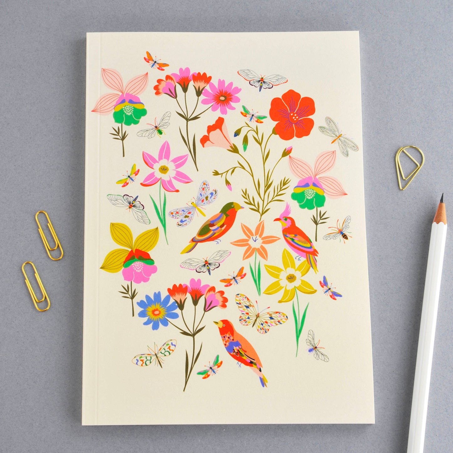 A cream-colored notebook with a colorful illustration of wildflowers, butterflies, and birds, styled on a gray background with gold paperclips and a white pencil.