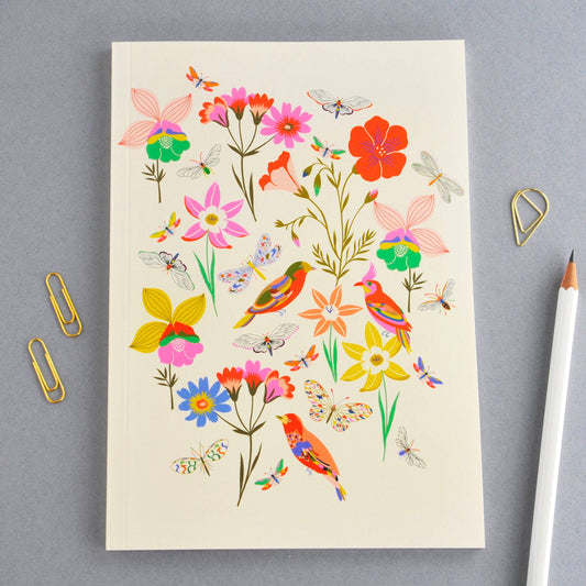 A cream-colored notebook with a colorful illustration of wildflowers, butterflies, and birds, styled on a gray background with gold paperclips and a white pencil.