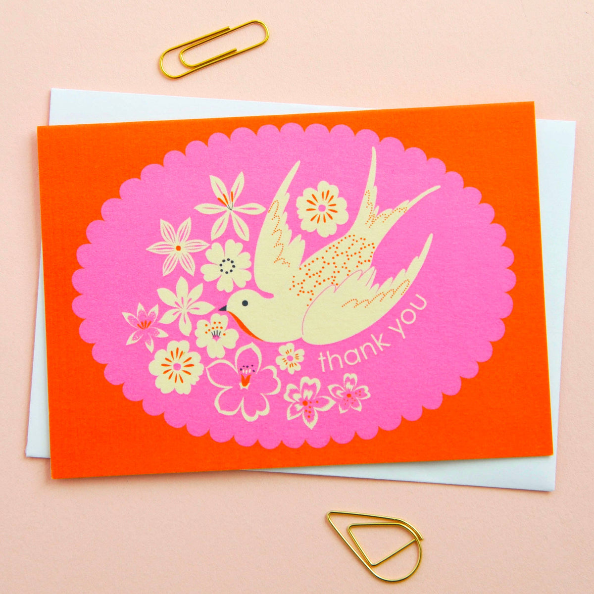 Greeting card with a bird and floral design on a pink background, placed on an orange card with gold paper clips.