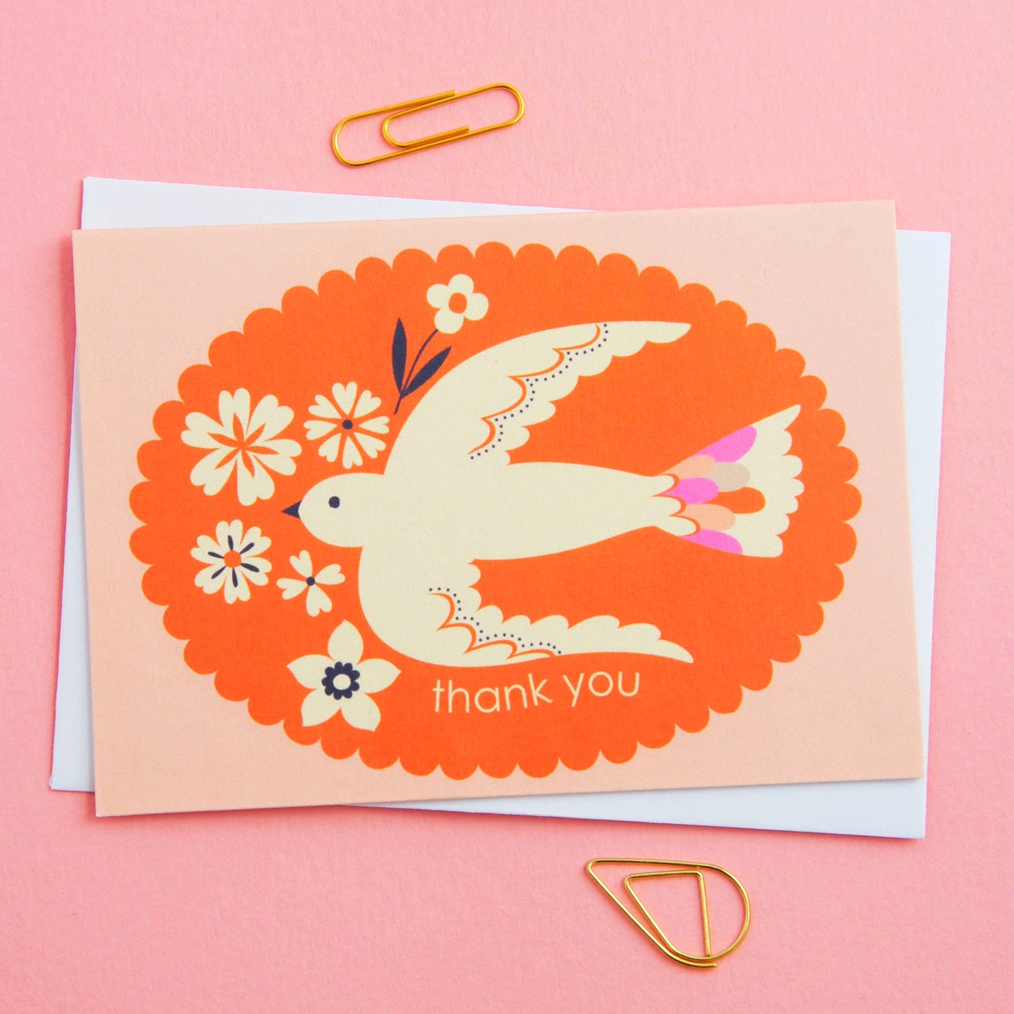 Greeting card with a dove and flowers on an orange background, placed on a pink surface with paper clips.