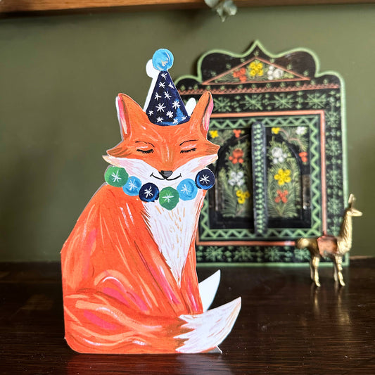 Decorative fox figurine with a party hat on a wooden surface with a decorative mirror in the background.