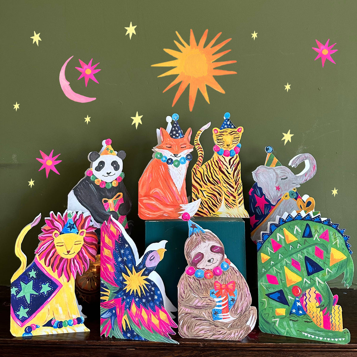 Colorful animal figurines with festive hats on a dark surface against a green background with sun and moon.