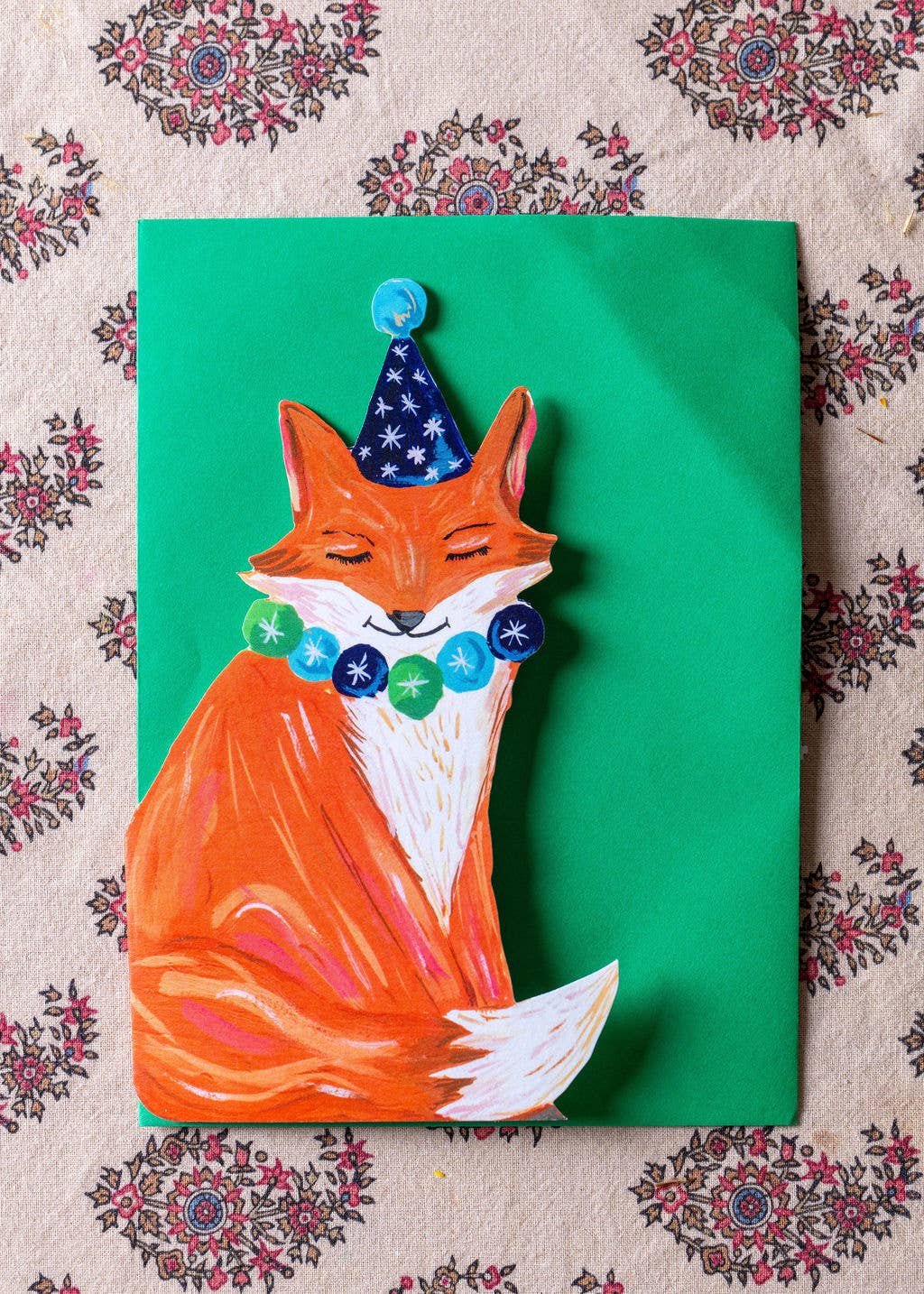 Painting of a fox wearing a party hat and beads on a green background with floral patterns.