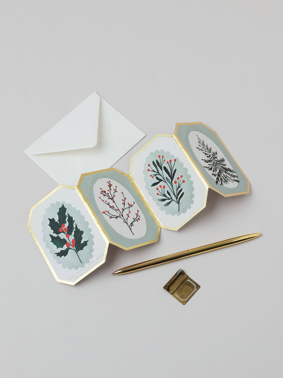 A set of four octagonal holiday greeting cards with gold borders, featuring botanical illustrations of holly, berries, and a fern on a light gray background. An open white envelope is positioned behind the cards, with a gold pen and a small gold square accessory placed in front.