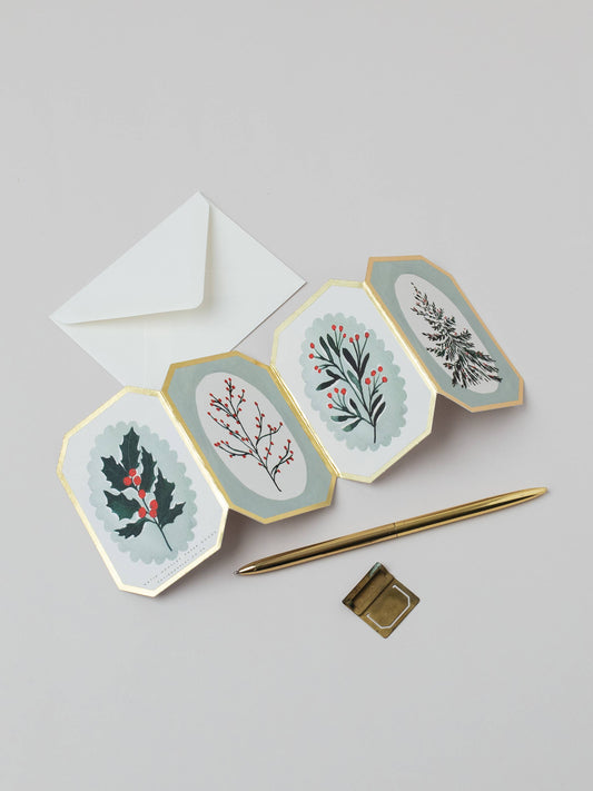 A set of four octagonal holiday greeting cards with gold borders, featuring botanical illustrations of holly, berries, and a fern on a light gray background. An open white envelope is positioned behind the cards, with a gold pen and a small gold square accessory placed in front.