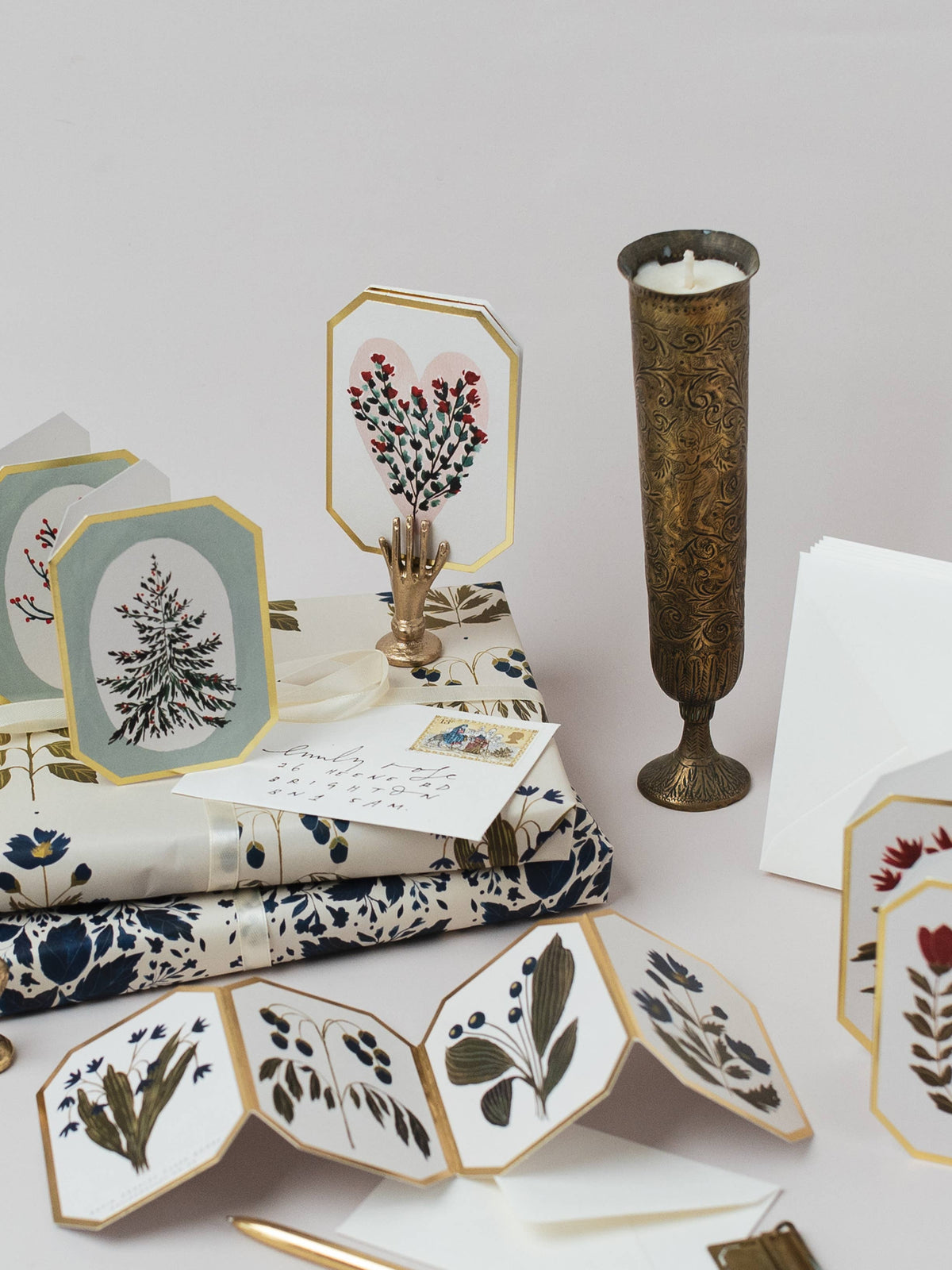 A styled flat-lay of coordinated stationery and home decor on a light neutral background, including patterned napkins or fabric pieces with botanical illustrations, octagonal cards or placemats with floral designs, a white envelope, and a tall ornate brass candle holder.