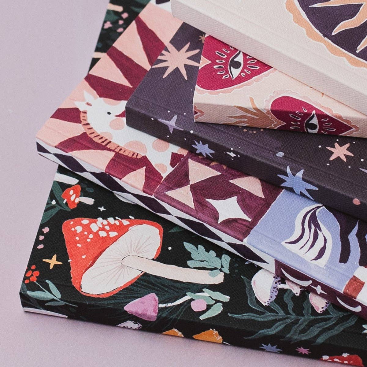 A stack of decorative notebooks with whimsical illustrated covers, including mushrooms, eyes, stars, and abstract shapes, photographed on a pale pink background.