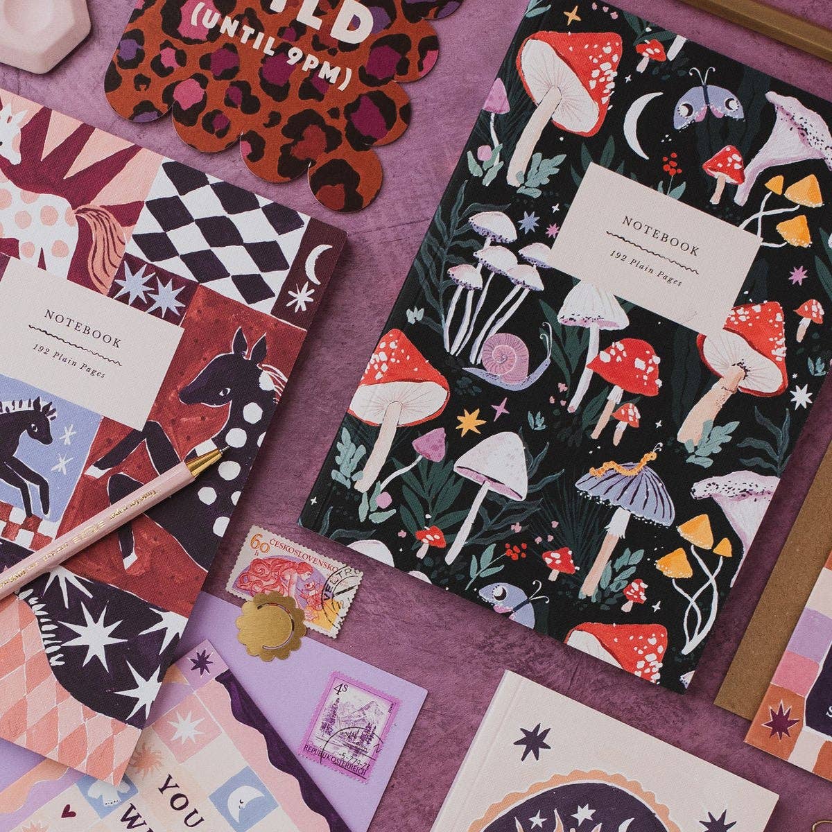 A styled flat-lay of multiple notebooks with whimsical illustrated covers, including mushrooms, stars, horses, and abstract patterns, arranged on a purple surface with a pen and small stationery pieces.