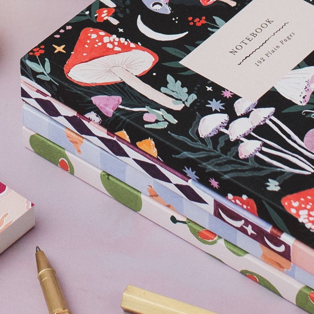 A stack of illustrated notebooks with a dark forest-themed cover featuring red and white mushrooms, white jellyfish, and a cream label reading 'NOTEBOOK 128 Flat Pages'. The notebooks are arranged on a light lavender surface with a gold pen and a cream pen visible nearby.