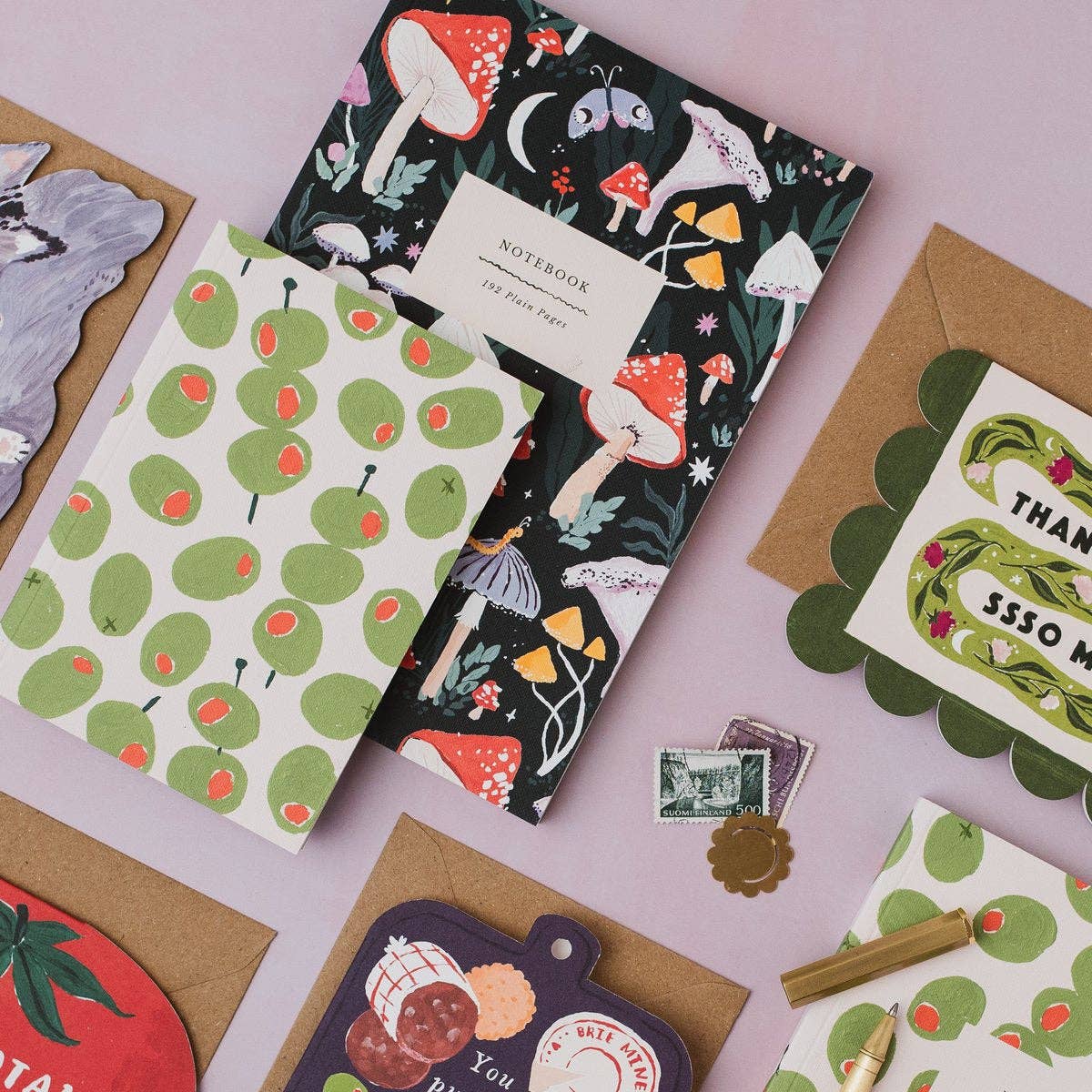 A styled flat-lay of paper goods on a light pink background, featuring a black notebook with colorful woodland mushroom and moon illustrations, a cream notebook with green pears, kraft envelopes, and matching printed cards and tags.