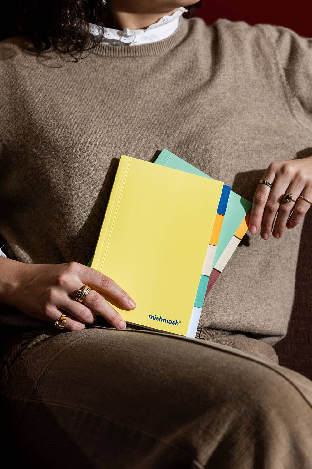 A person in a beige sweater holds a stack of colorful notebooks or planners, with the top one in bright yellow showing the 'mishmash' logo near the bottom.