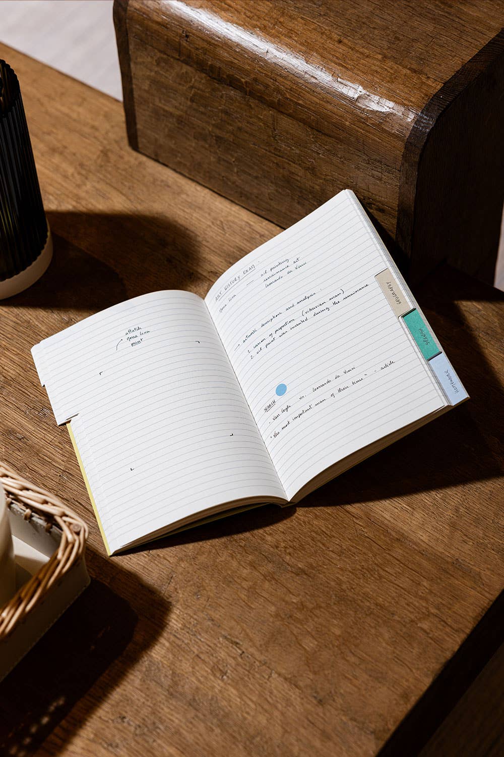 An open lined notebook with handwritten notes and colorful page tabs, placed on a wooden table in warm natural light.