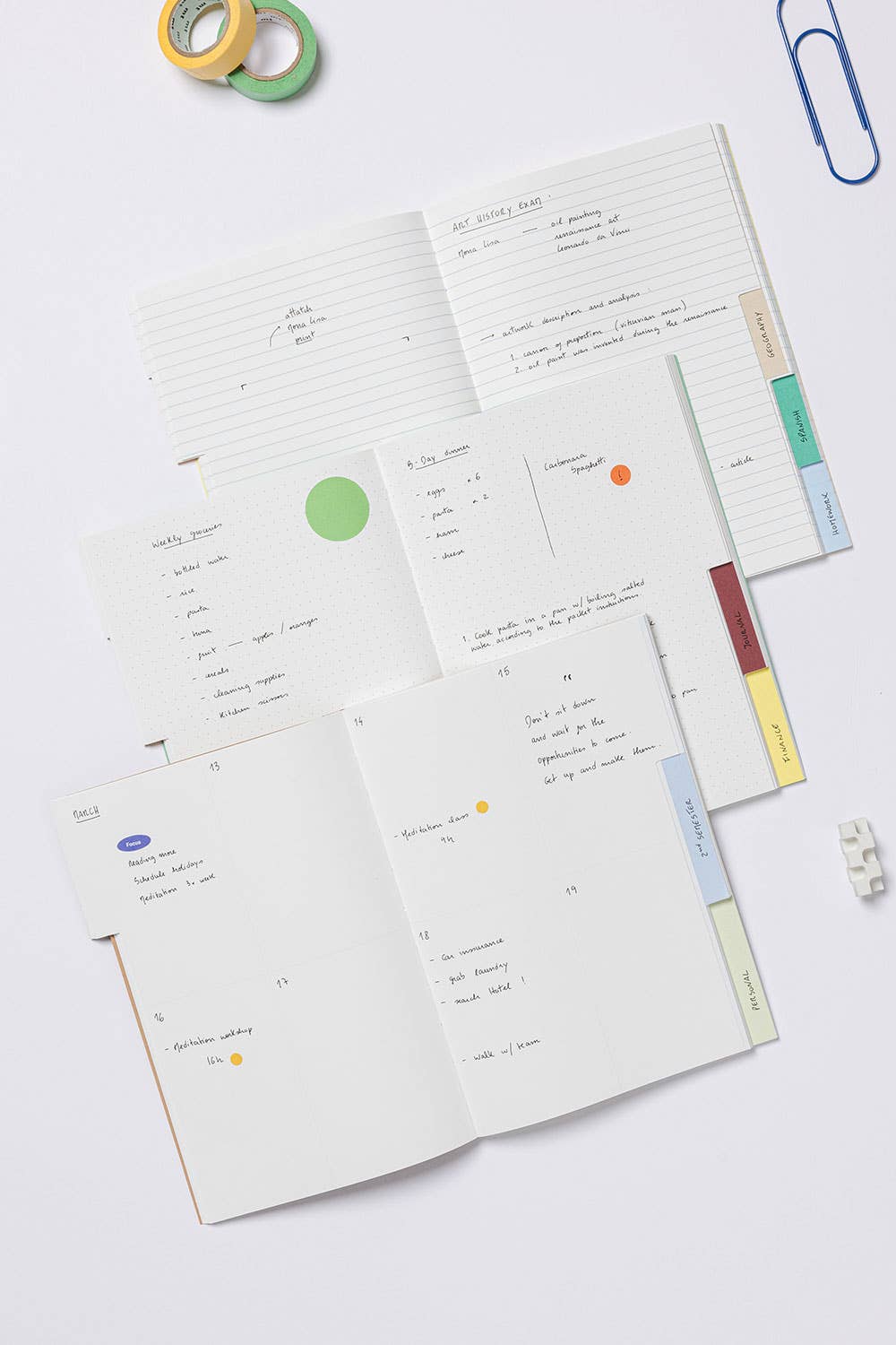 An open planner or notebook spread on a light gray background, showing multiple interior pages with handwritten notes, colored page dividers, sticky notes, and a few stationery props around it.