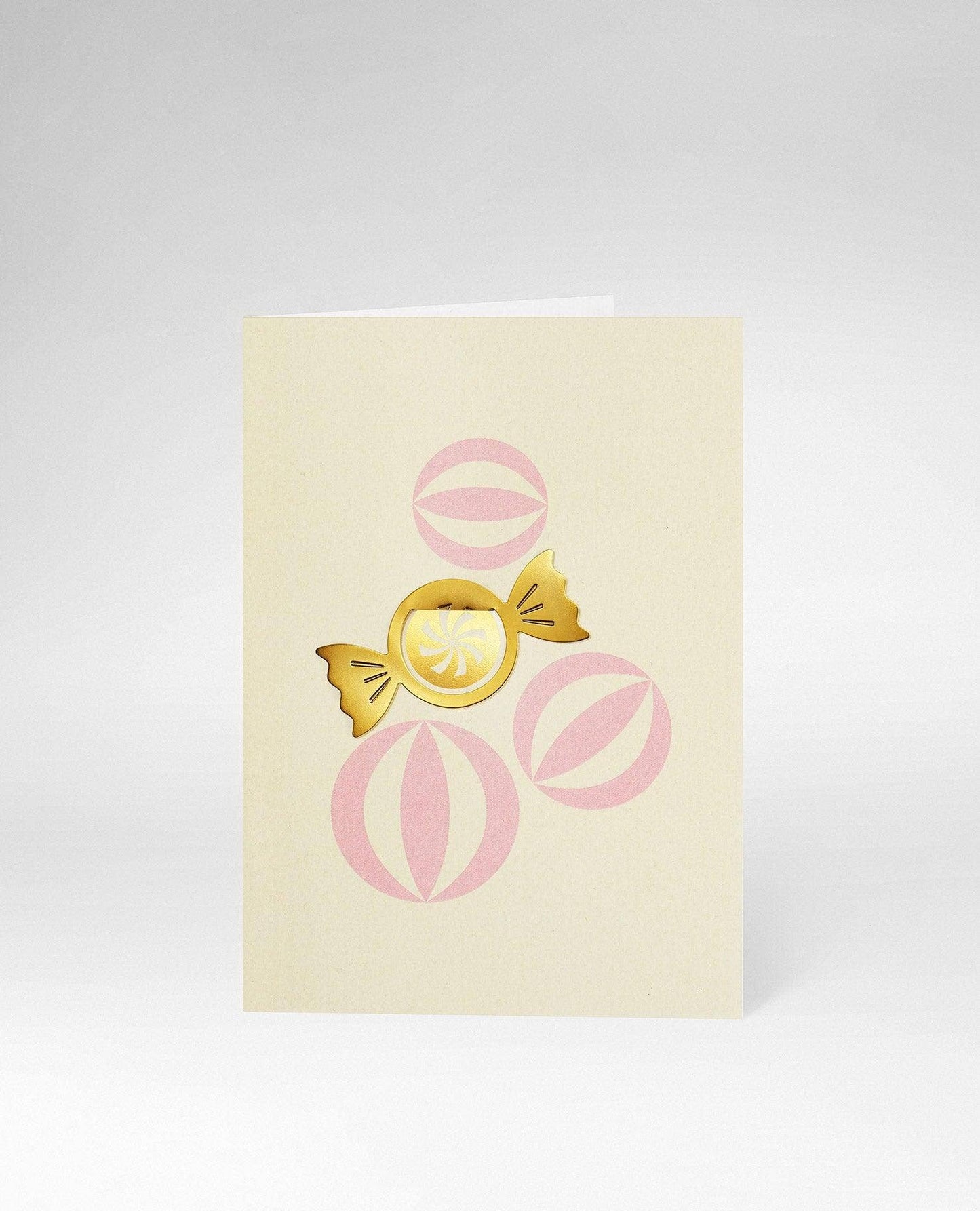 A cream-colored greeting card featuring a central gold foil candy illustration with pink circular accents, photographed on a light gray background.