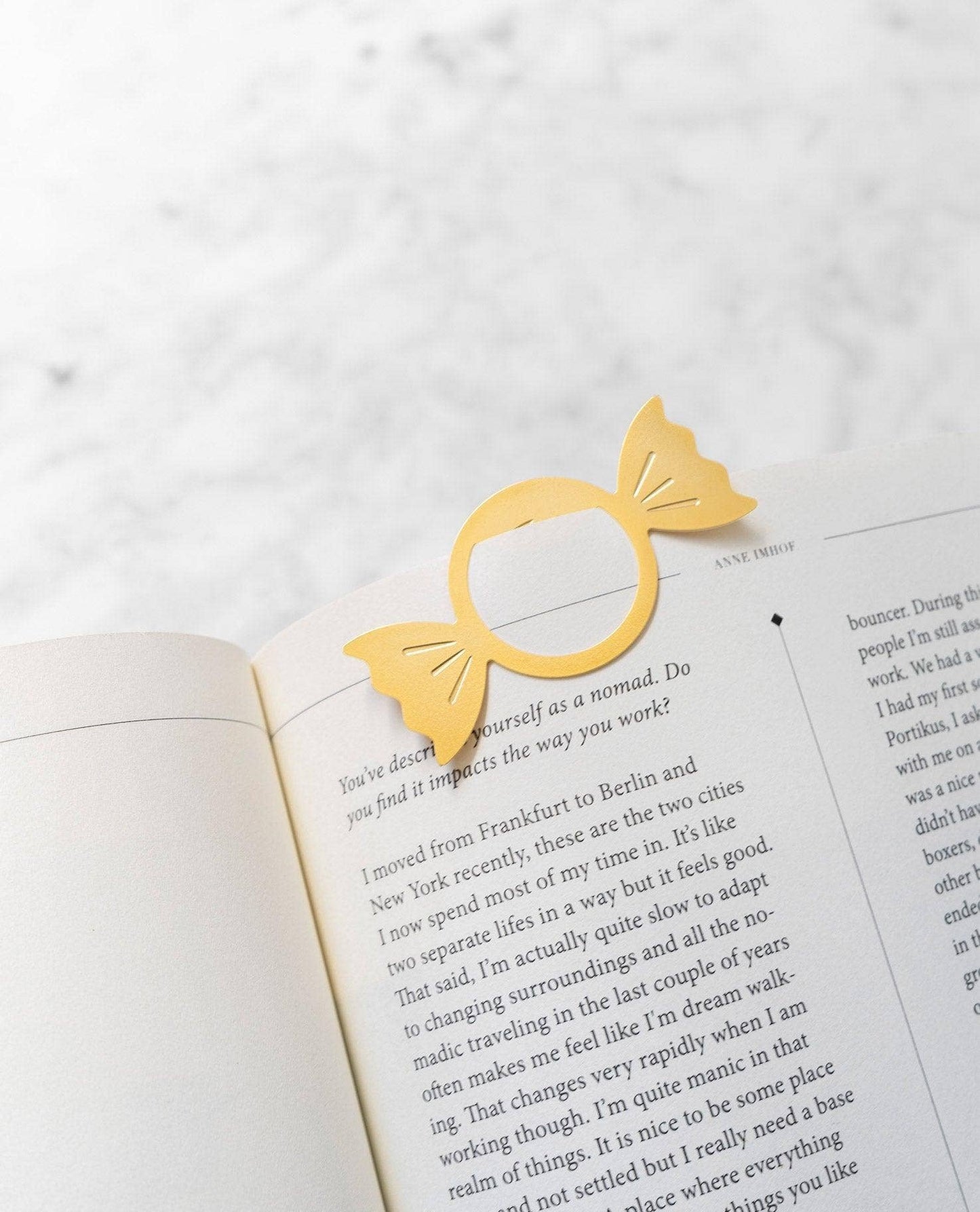 A gold paper bookmark shaped like a bow tie is placed on an open book page, photographed from above on a light marble-like surface.