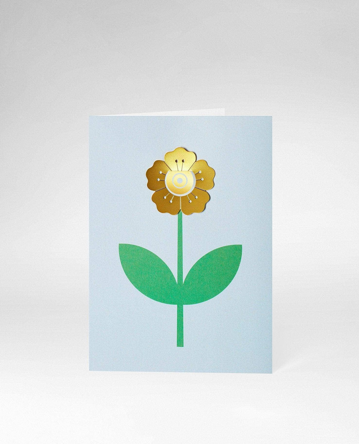 A light blue greeting card featuring a stylized gold and white flower with green leaves and stem, shown on a neutral light gray background.