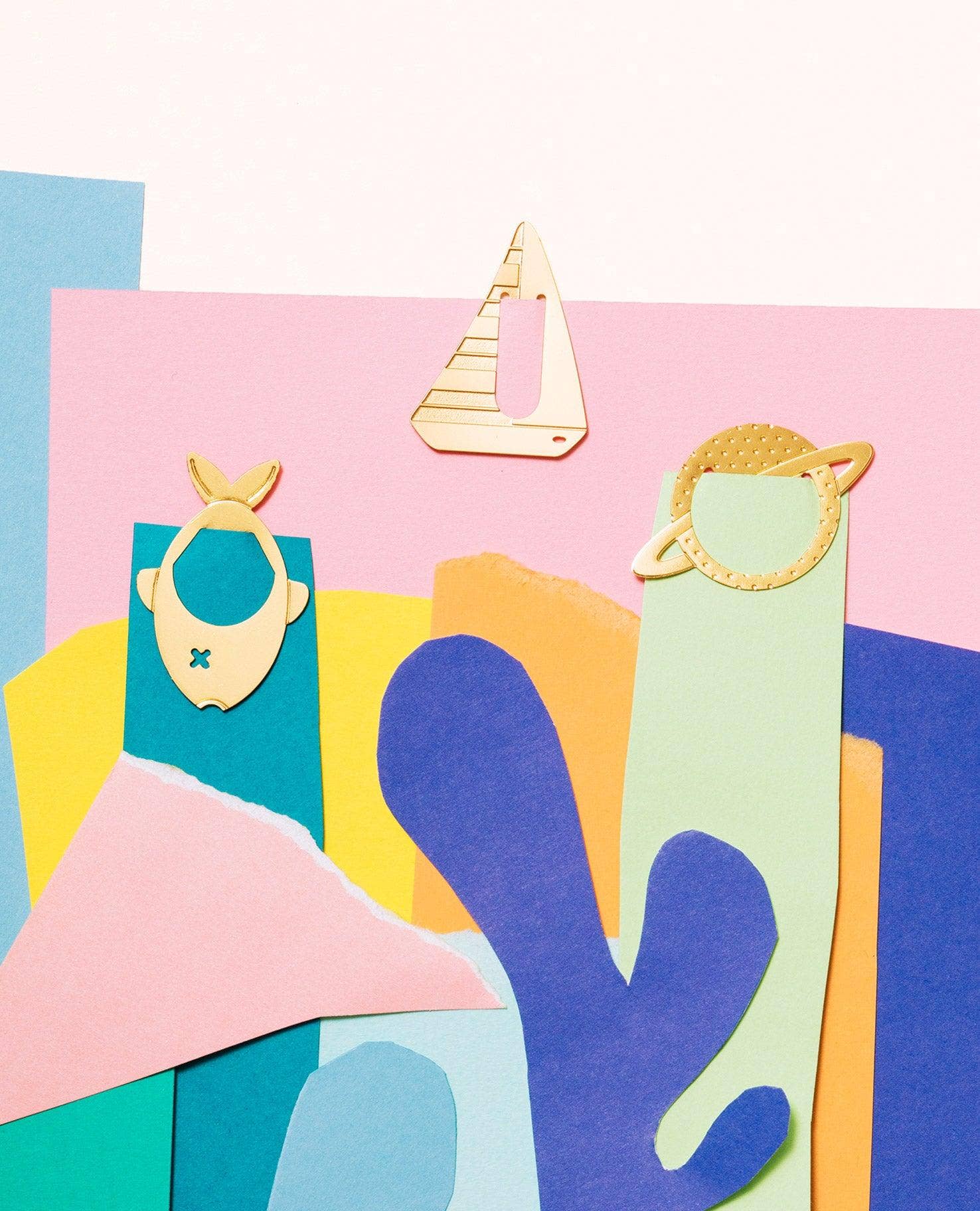 A colorful abstract collage of layered paper shapes in pink, blue, teal, yellow, and purple, with three small gold-tone decorative cutout figures placed on top.