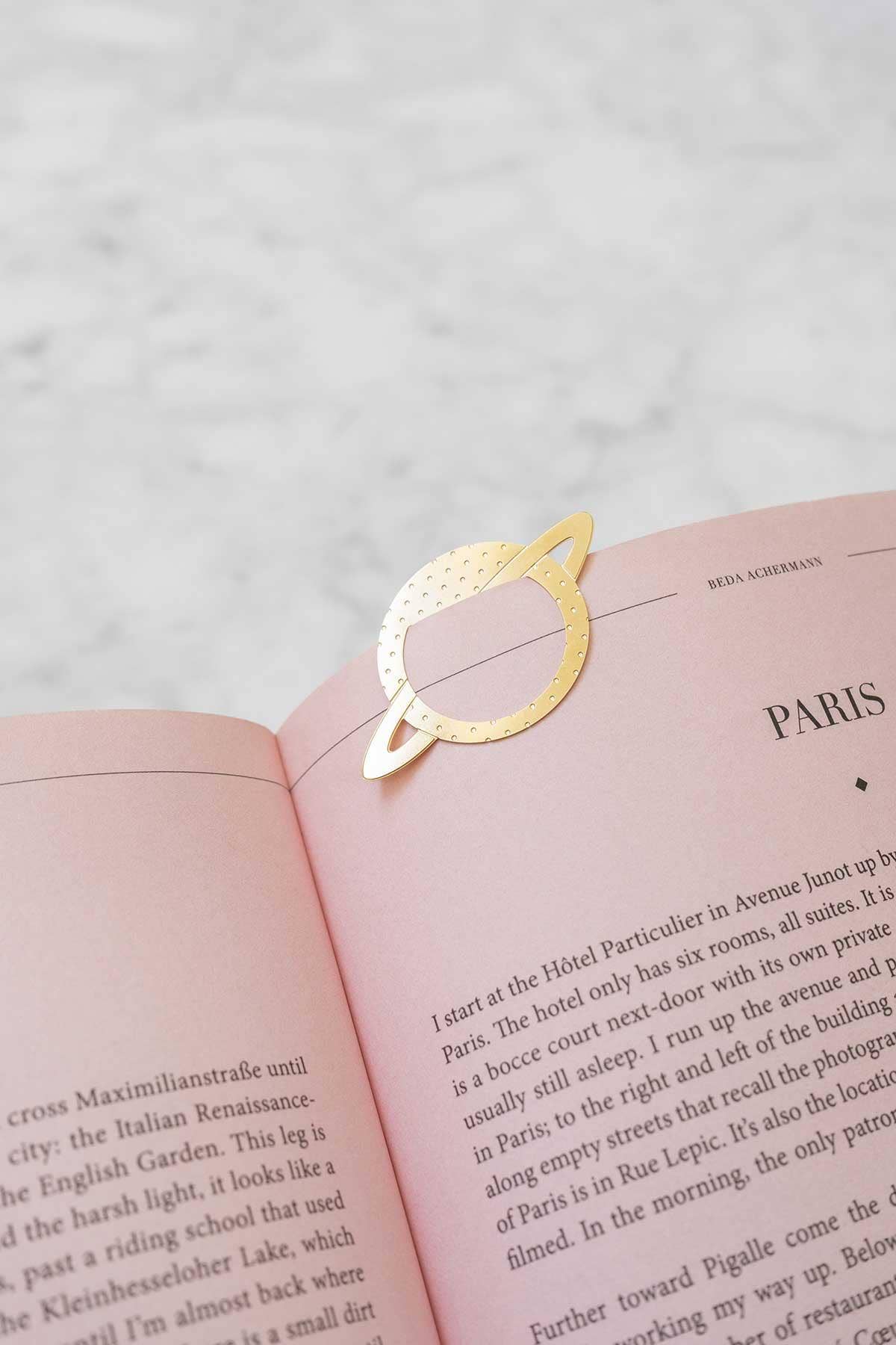 A gold metal Saturn-shaped bookmark with a dotted ring design rests on an open pink book, photographed on a light marble surface.