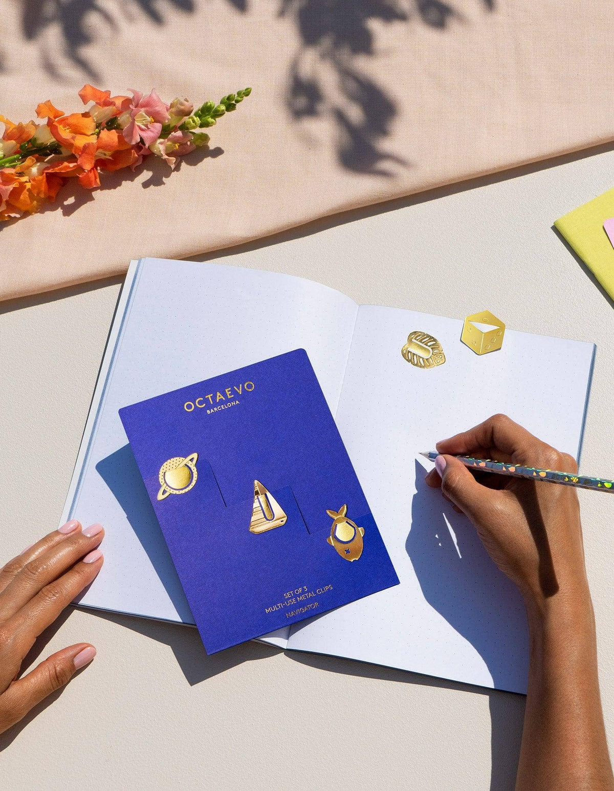 A person's hands are shown holding an open book, with a blue Octaveo card displaying three gold earrings (a ring, a boat, and a deer) on the left page. The right page has a small gold ring and a gold cube, with a beige surface, flowers, and a pen visible.