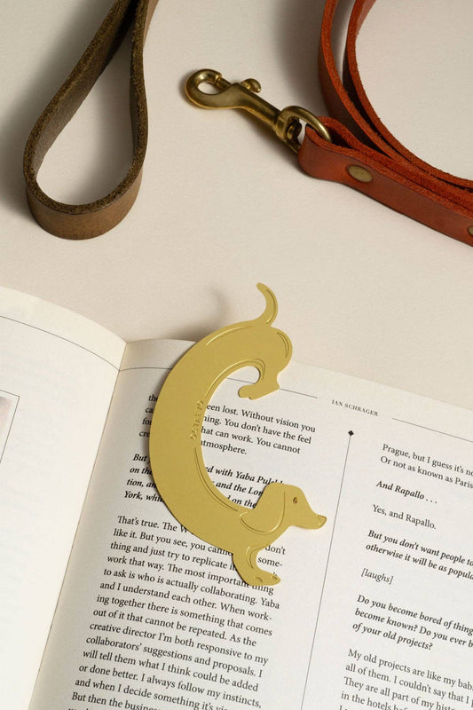A gold metal dog-shaped bookmark resting on an open book, with a brown leather strap and brass hardware placed above it on a white surface.