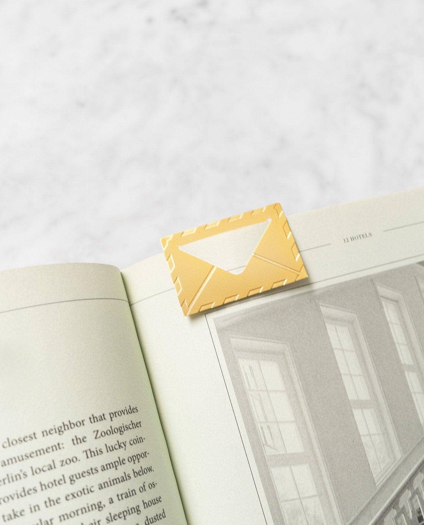 A gold envelope-shaped bookmark clipped to an open book on a light marble surface.