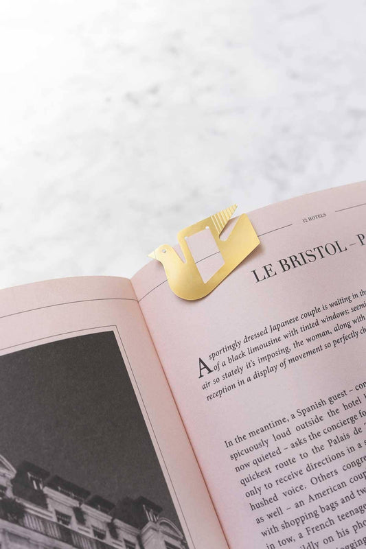 A gold metal bookmark shaped like a stylized coffee cup rests on an open book with pale pink pages, photographed on a light marble-like surface.