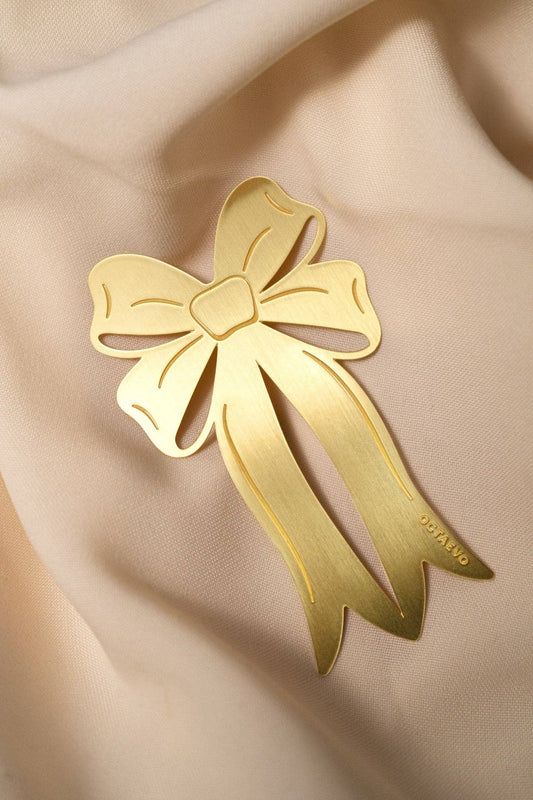 A gold bow-shaped bookmark with cutout details, resting on a draped beige fabric background.