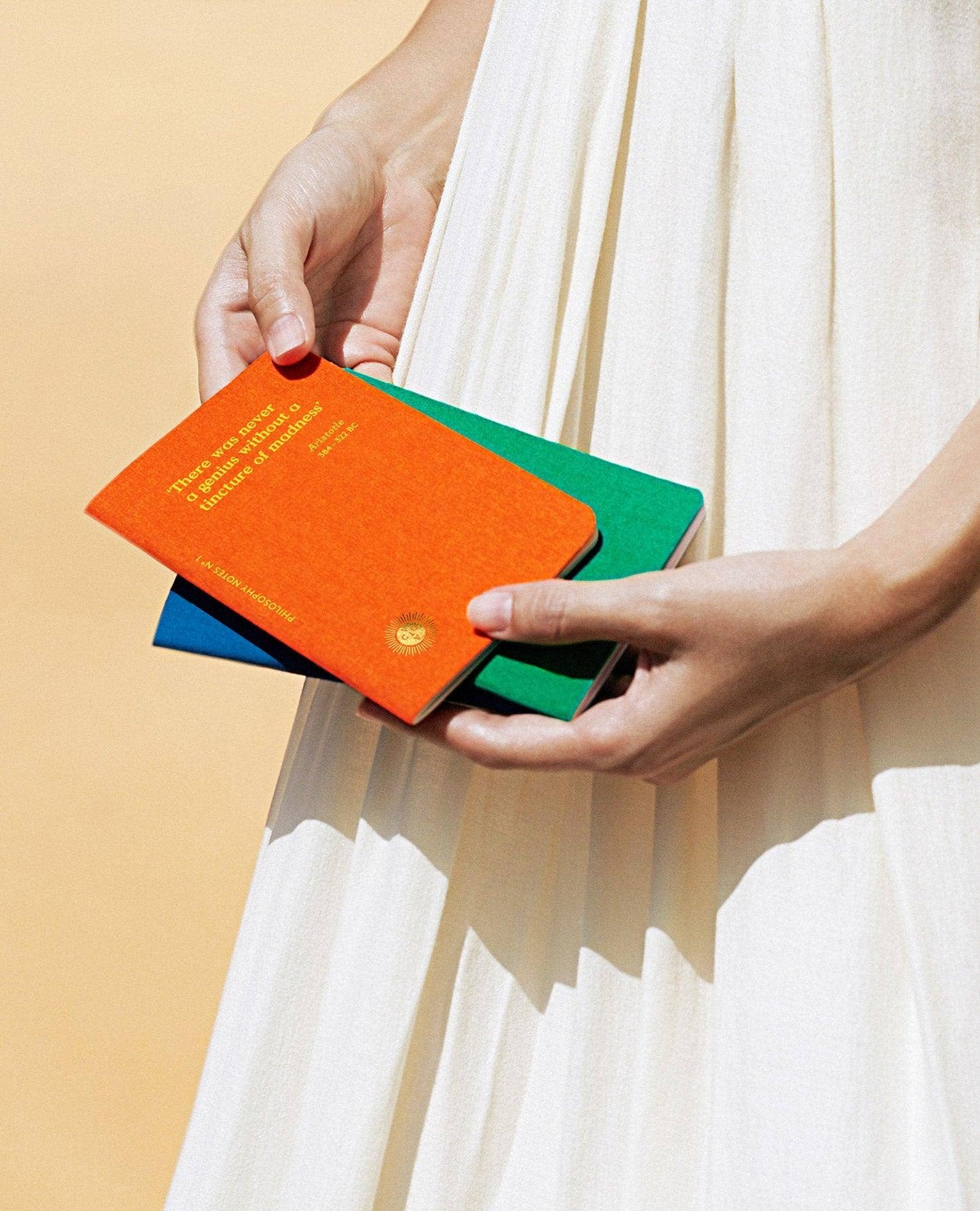 A person in a cream pleated dress holds three textured hardcover notebooks in orange, green, and blue, with the orange notebook on top featuring gold text.