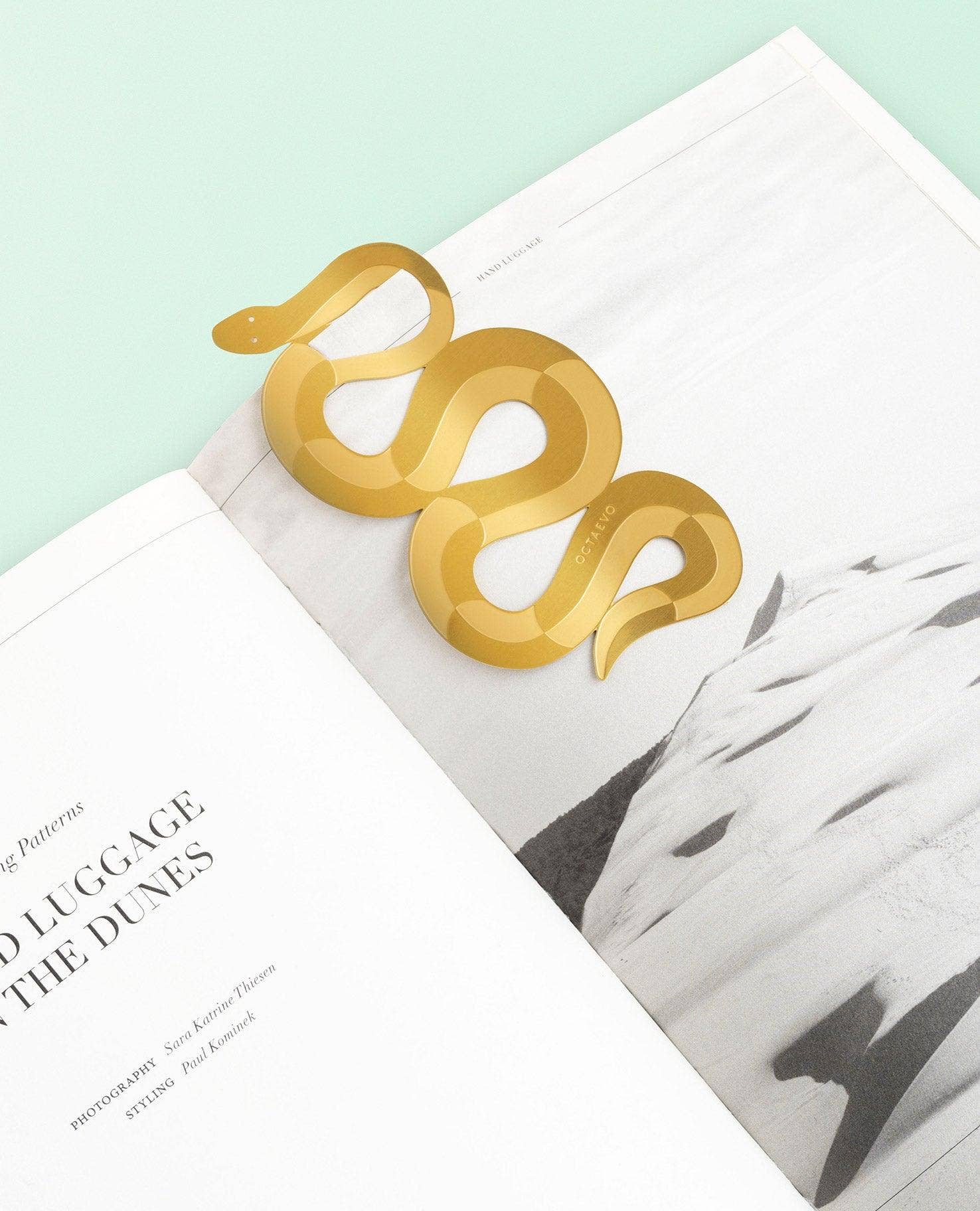 A metallic gold snake-shaped bookmark resting on open magazine pages against a pale mint background.